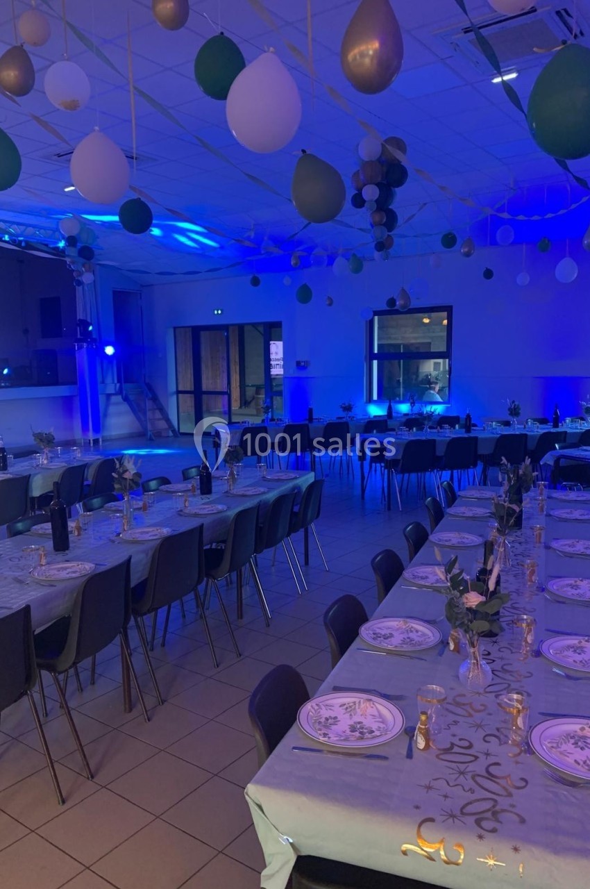 Salle décorée pour un événement, avec tables dressées, ballons suspendus et éclairage bleu tamisé.