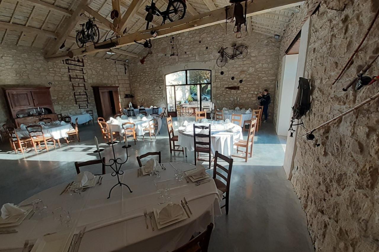 Salle en pierre avec tables dressées pour un repas, éclairée par la lumière naturelle entrant par une grande baie vitrée.