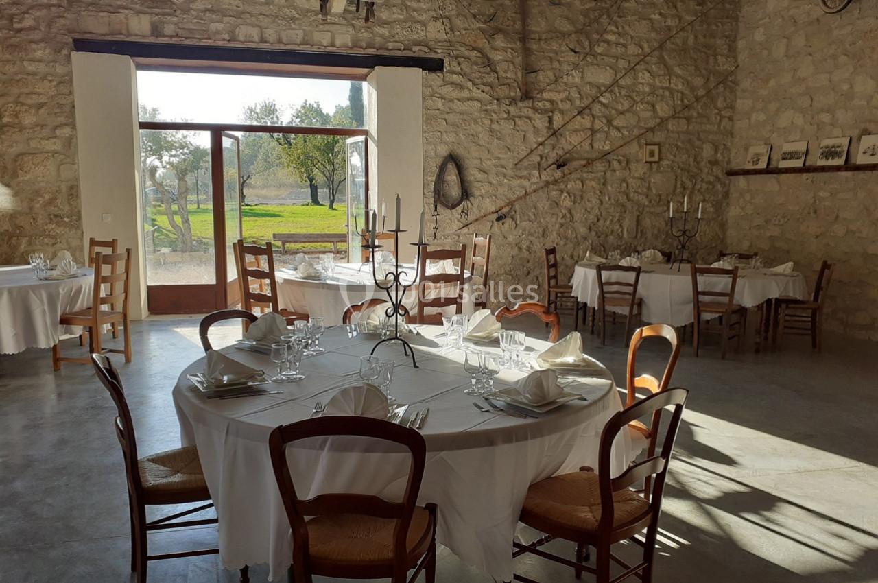 Salle de réception lumineuse avec tables rondes dressées, nappes blanches et vue sur un jardin à travers une porte vitrée.