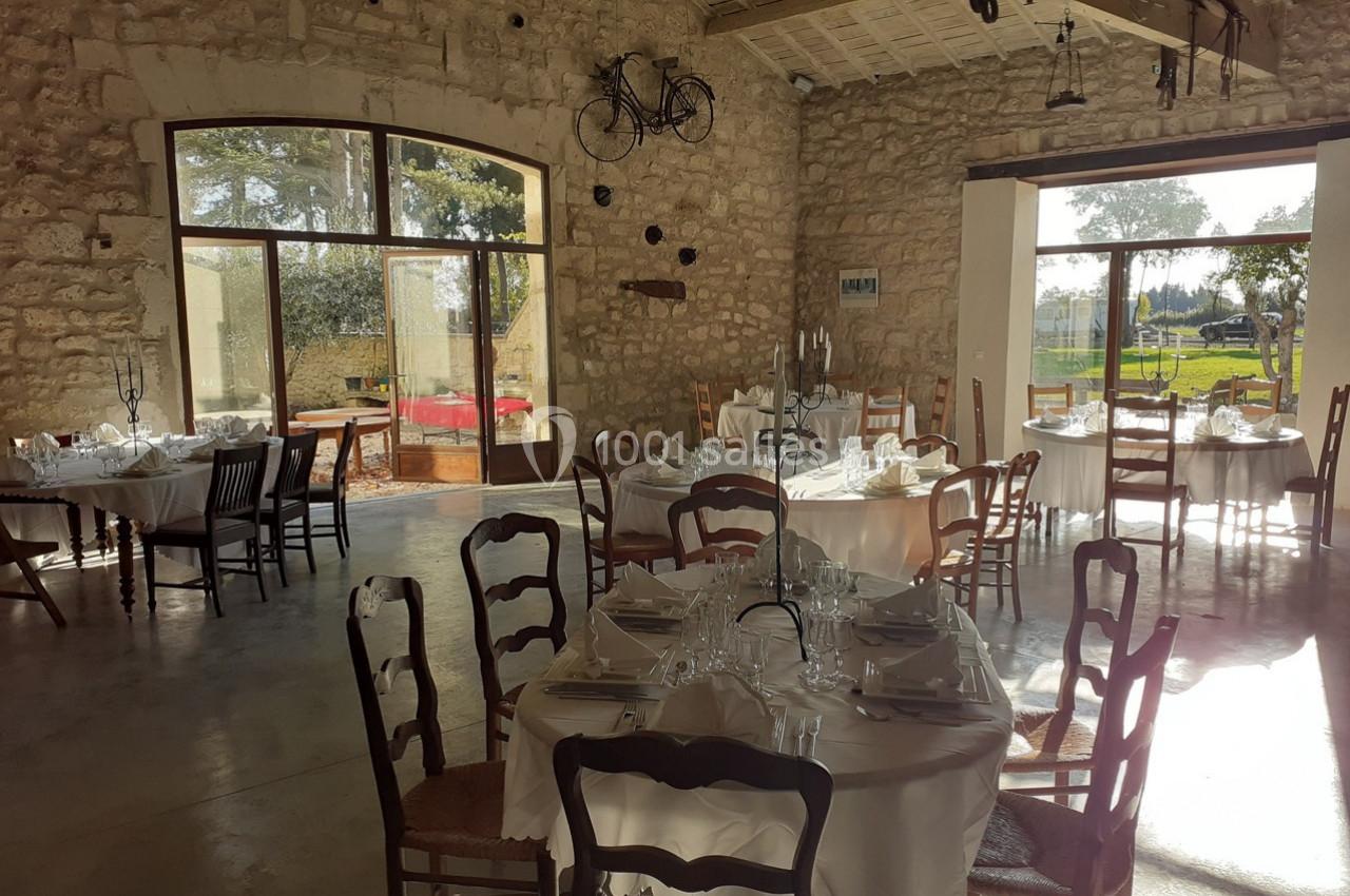 Salle de réception lumineuse avec des tables rondes dressées, des chaises en bois et des murs en pierre apparente.