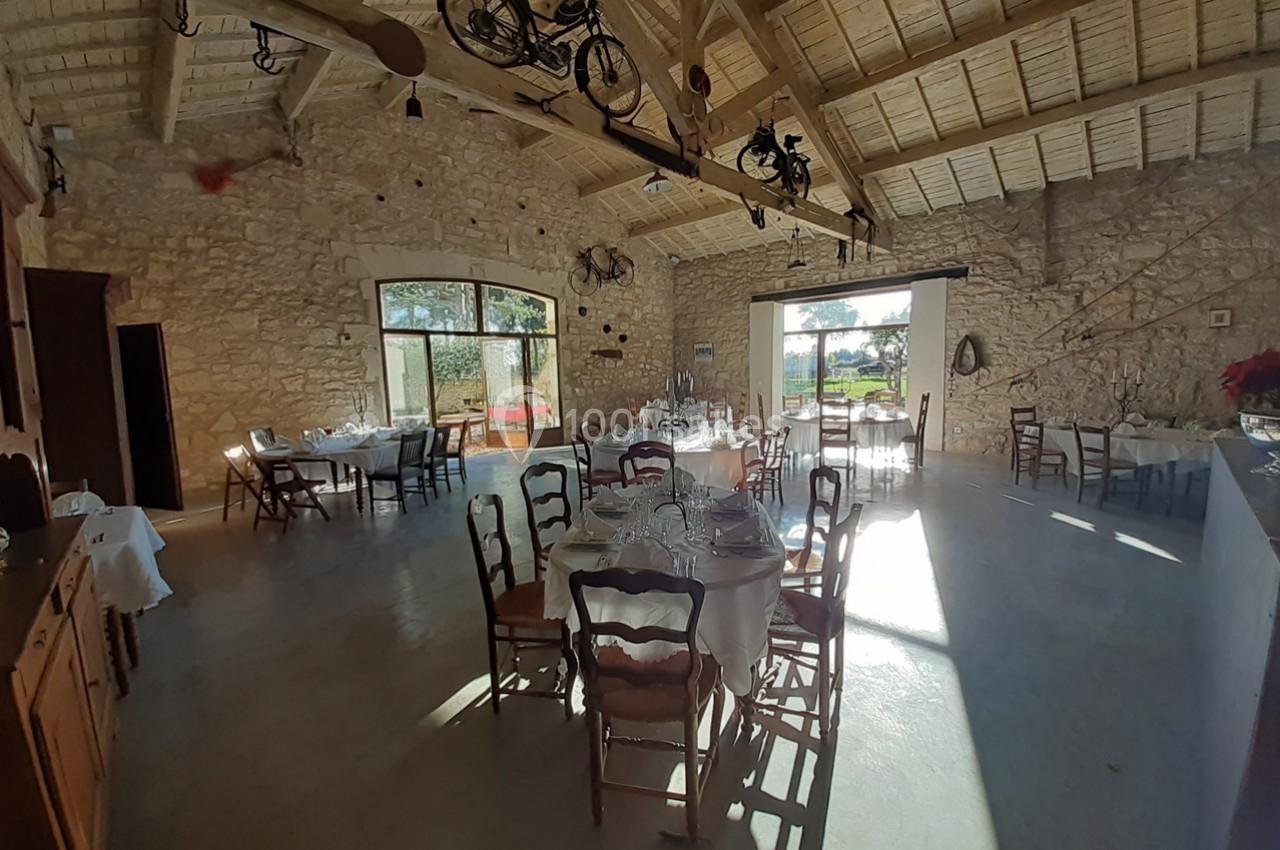 Salle lumineuse avec murs en pierre, tables dressées et décorations suspendues, donnant sur un extérieur ensoleillé.