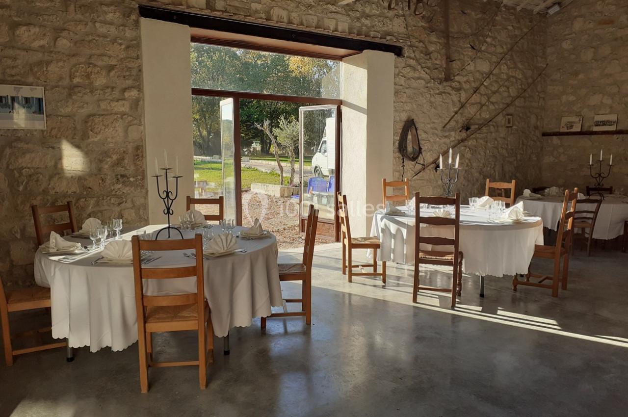 Salle en pierre lumineuse avec tables rondes dressées, nappes blanches et chaises en bois, donnant sur un extérieur…