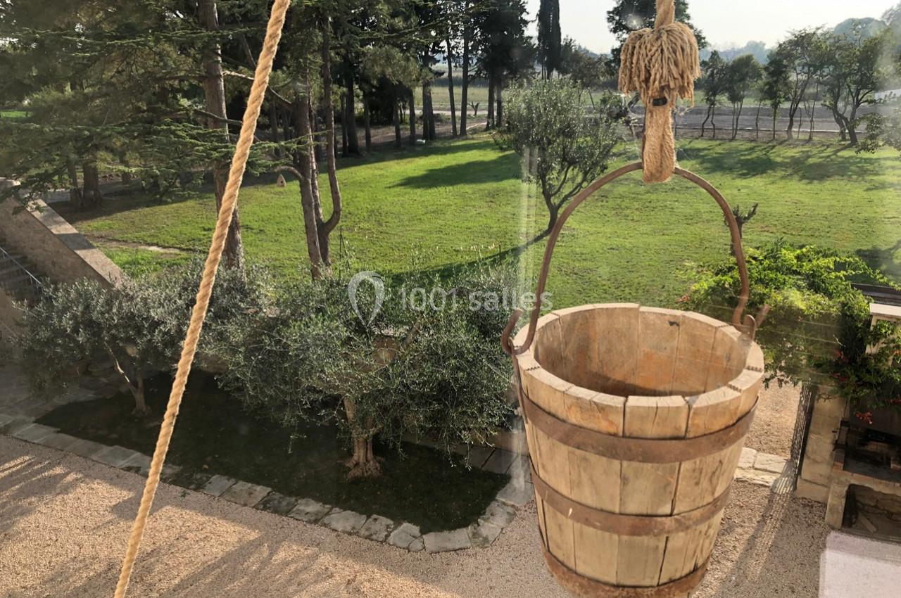 Seau en bois suspendu par une corde, avec vue sur un jardin verdoyant et des arbres en arrière-plan.