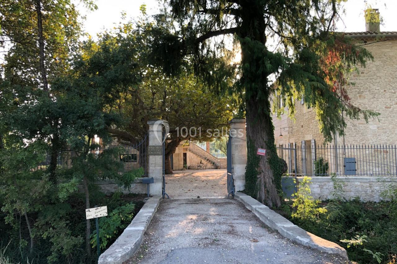 Entrée d'une propriété avec portail ouvert, allée bordée d'arbres et bâtiment en pierre en arrière-plan.
