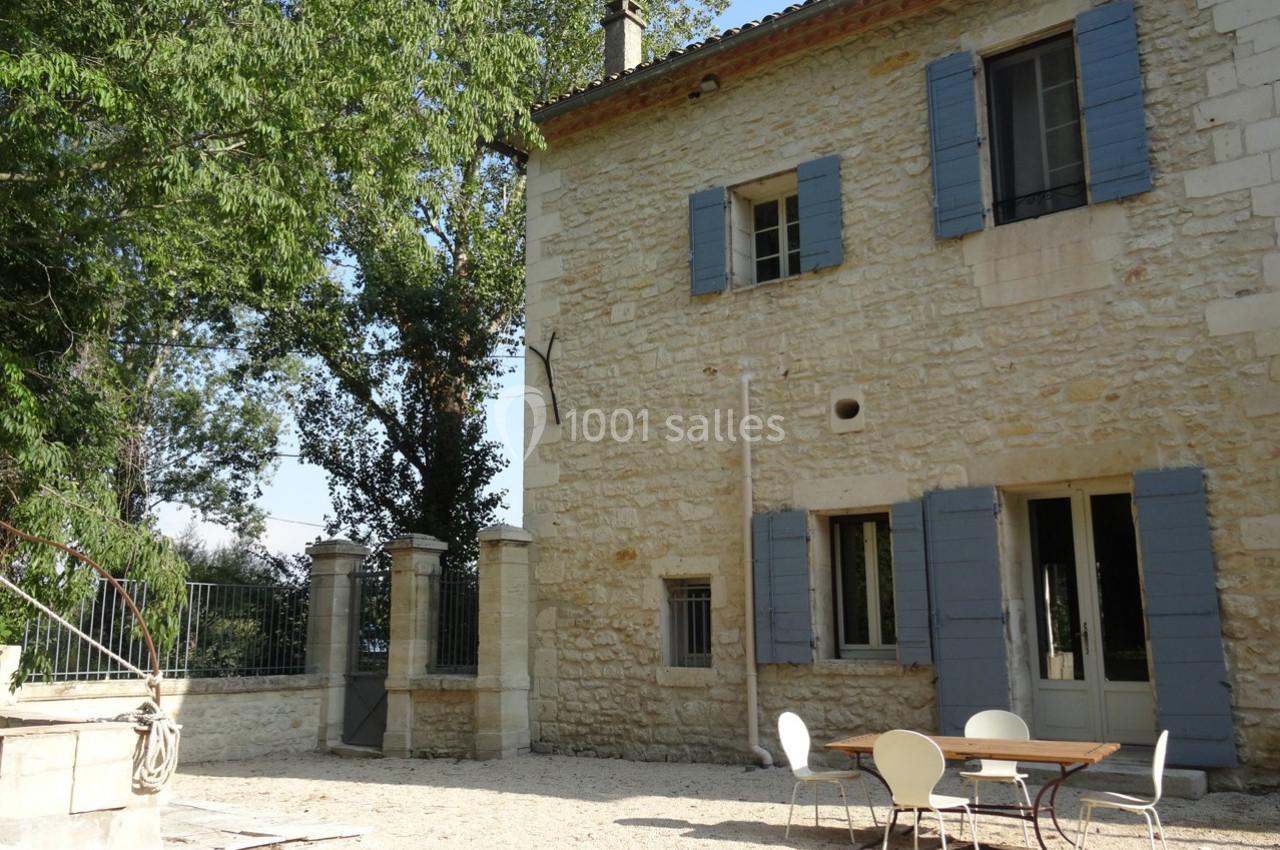 Façade en pierre d'une maison avec volets bleus, table et chaises sur une cour en gravier, entourée de végétation.