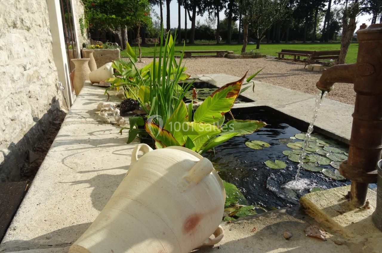 Fontaine ancienne avec pompe à eau, bassin orné de nénuphars et plantes, entouré de pots en terre cuite et jardin verdoyant.