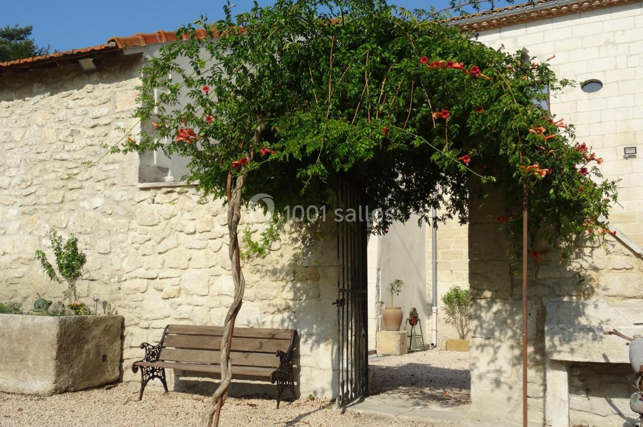 Arche végétale fleurie devant une maison en pierre, avec un banc en bois et un sol gravillonné.