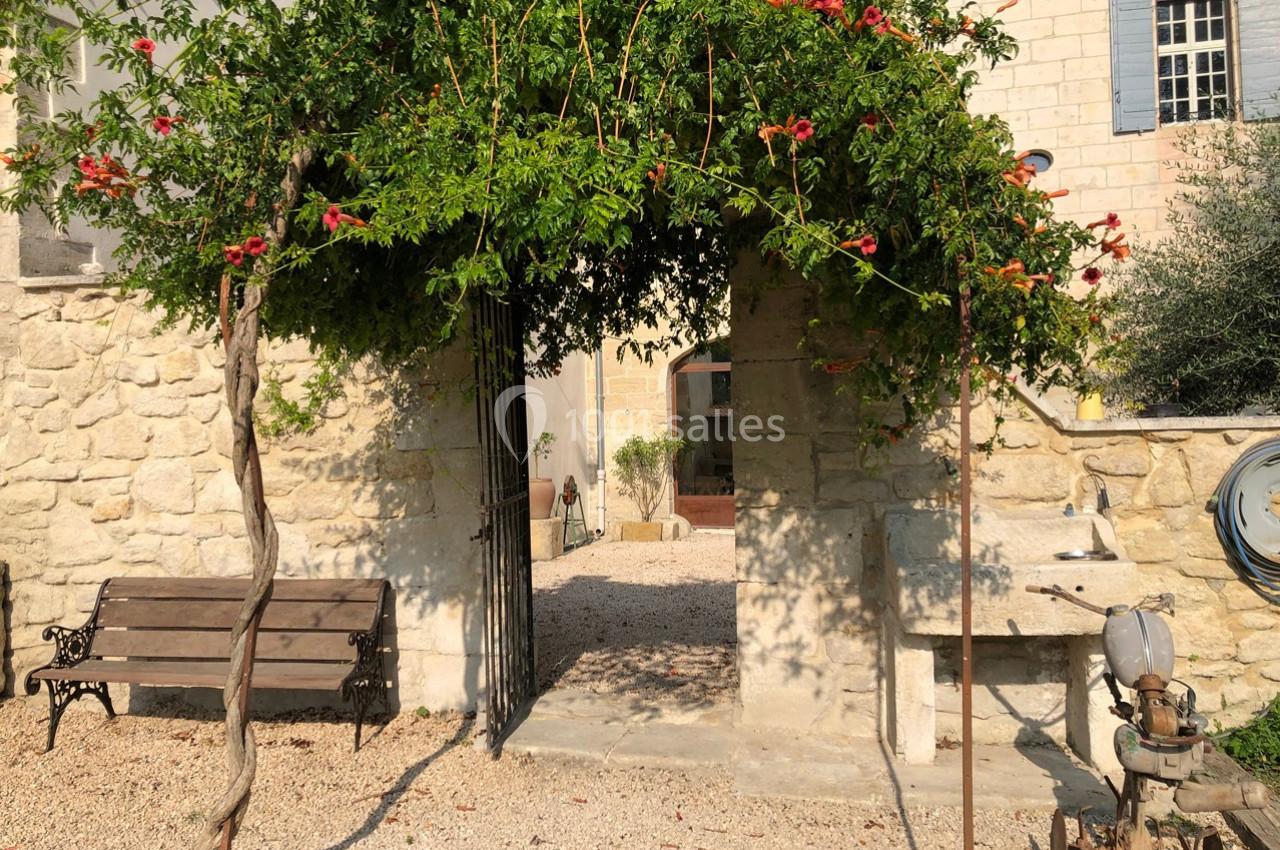 Arche en pierre recouverte de végétation fleurie, donnant sur une cour en gravier avec un banc et des éléments en pierre.
