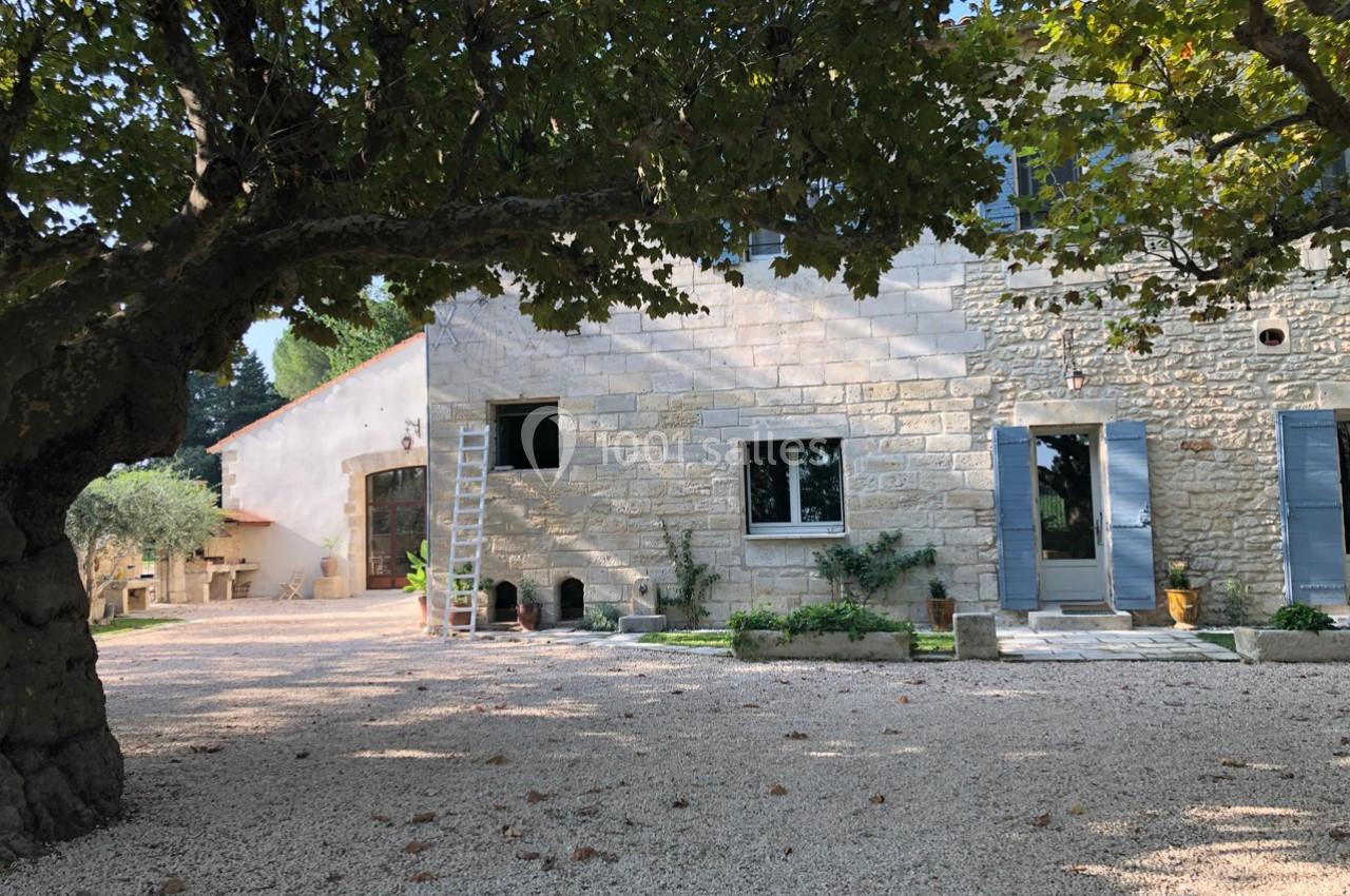 Façade en pierre d'une maison avec volets bleus, entourée d'arbres et d'une cour en gravier.