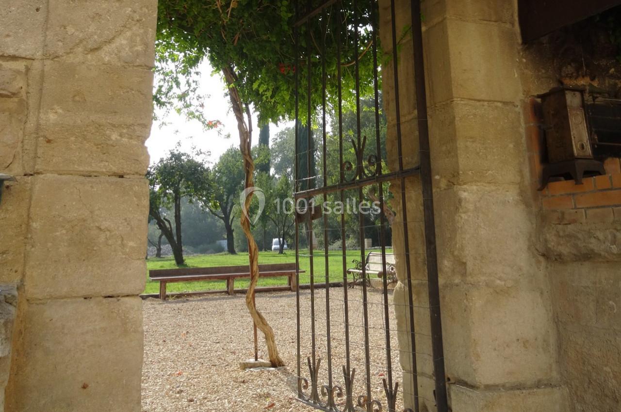 Portail en fer forgé ouvert sur un jardin avec bancs, arbres et lumière naturelle filtrée par les feuillages.