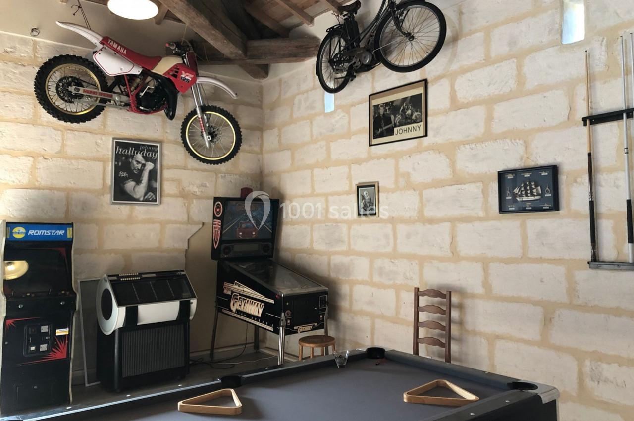 Salle de jeux avec billard, flipper, bornes d'arcade, vélos et moto accrochés au mur en pierre apparente.