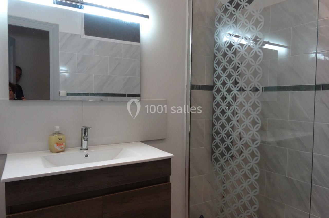 Salle de bain moderne avec lavabo, meuble en bois sombre, miroir éclairé et douche avec paroi en verre décorée.