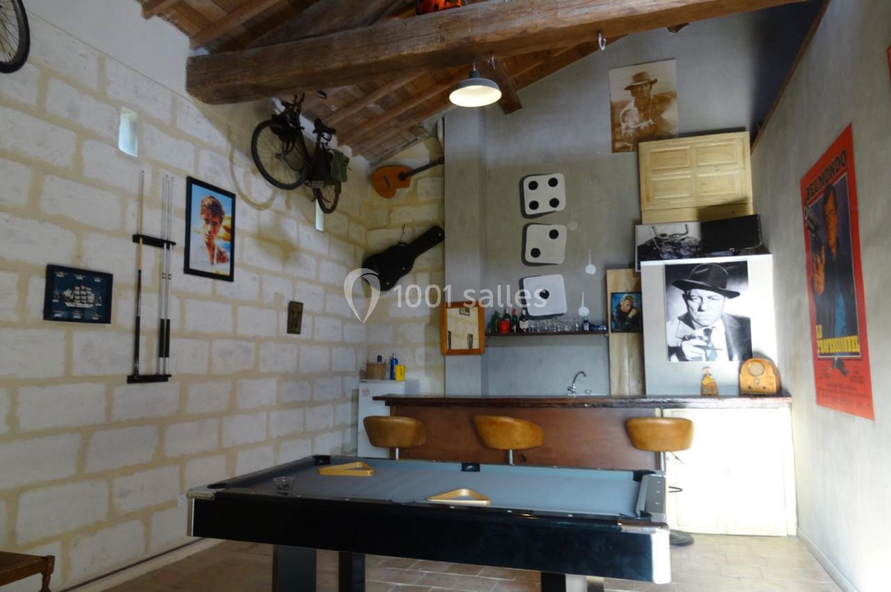 Salle de loisirs avec billard, bar, décorations murales variées et plafond en poutres apparentes.