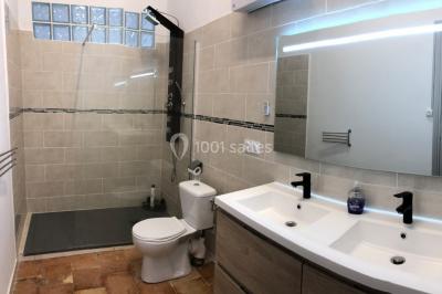 Salle de bain avec douche à l'italienne, lavabo double vasque, miroir éclairé et toilettes sur carrelage en terre cuite.