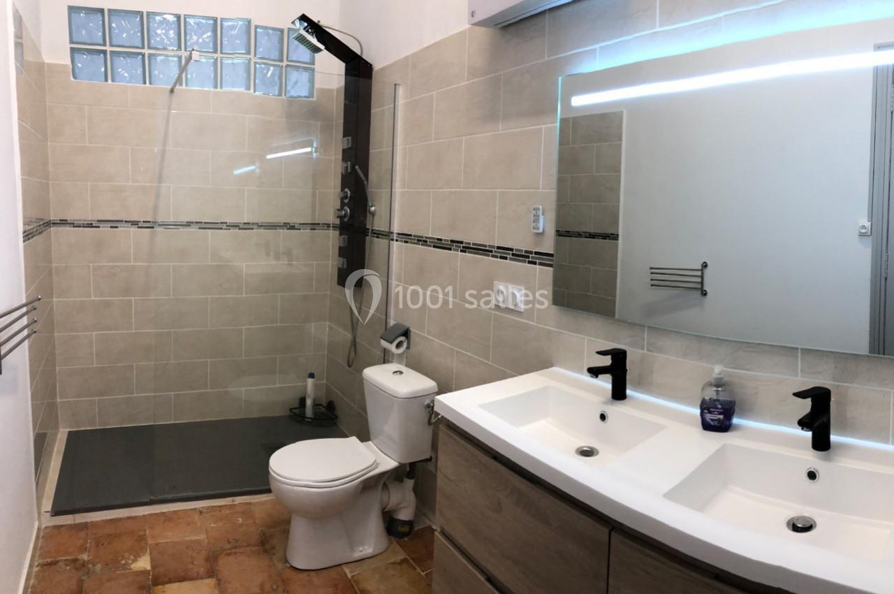 Salle de bain avec douche à l'italienne, lavabo double vasque, miroir éclairé et toilettes sur carrelage en terre cuite.