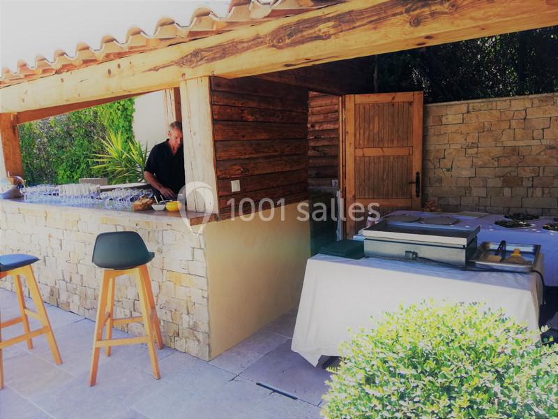 Un homme prépare des plats dans une cuisine extérieure en bois et pierre, avec un comptoir et des tables aménagées.