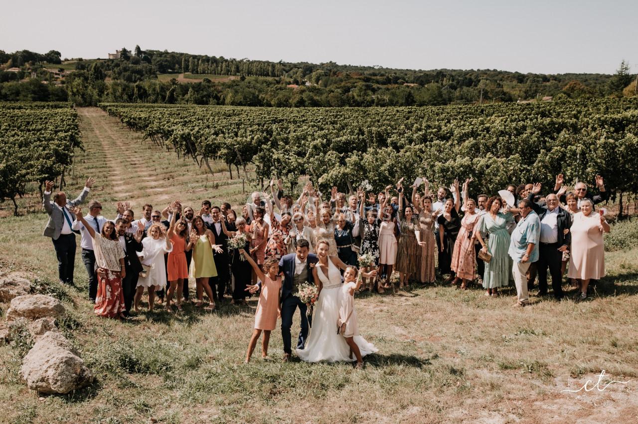 Un groupe de personnes pose dans un vignoble sous un ciel dégagé, avec un couple en tenue de mariage au premier plan.