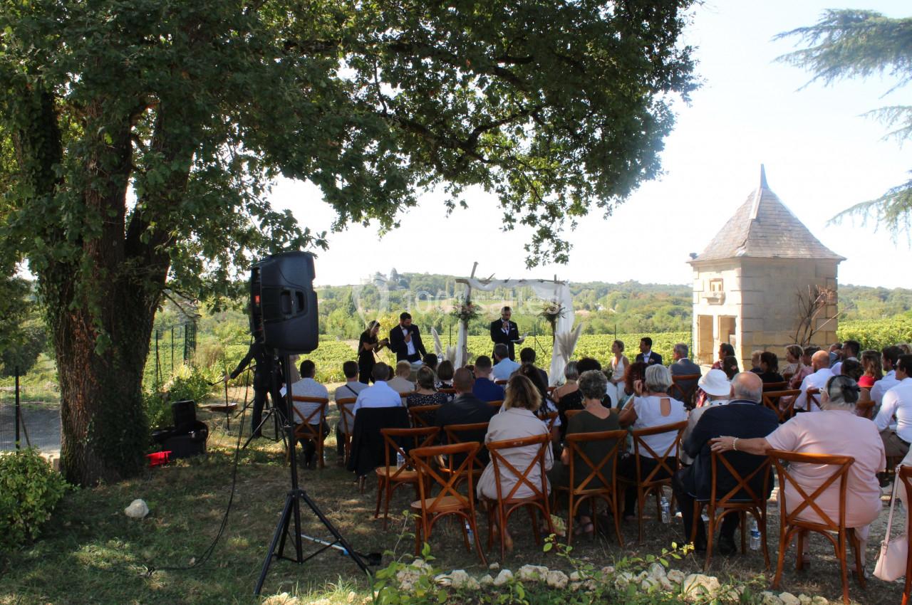 Cérémonie en plein air avec des invités assis, face à un officiant et un couple, entourés de verdure et d'un bâtiment en…