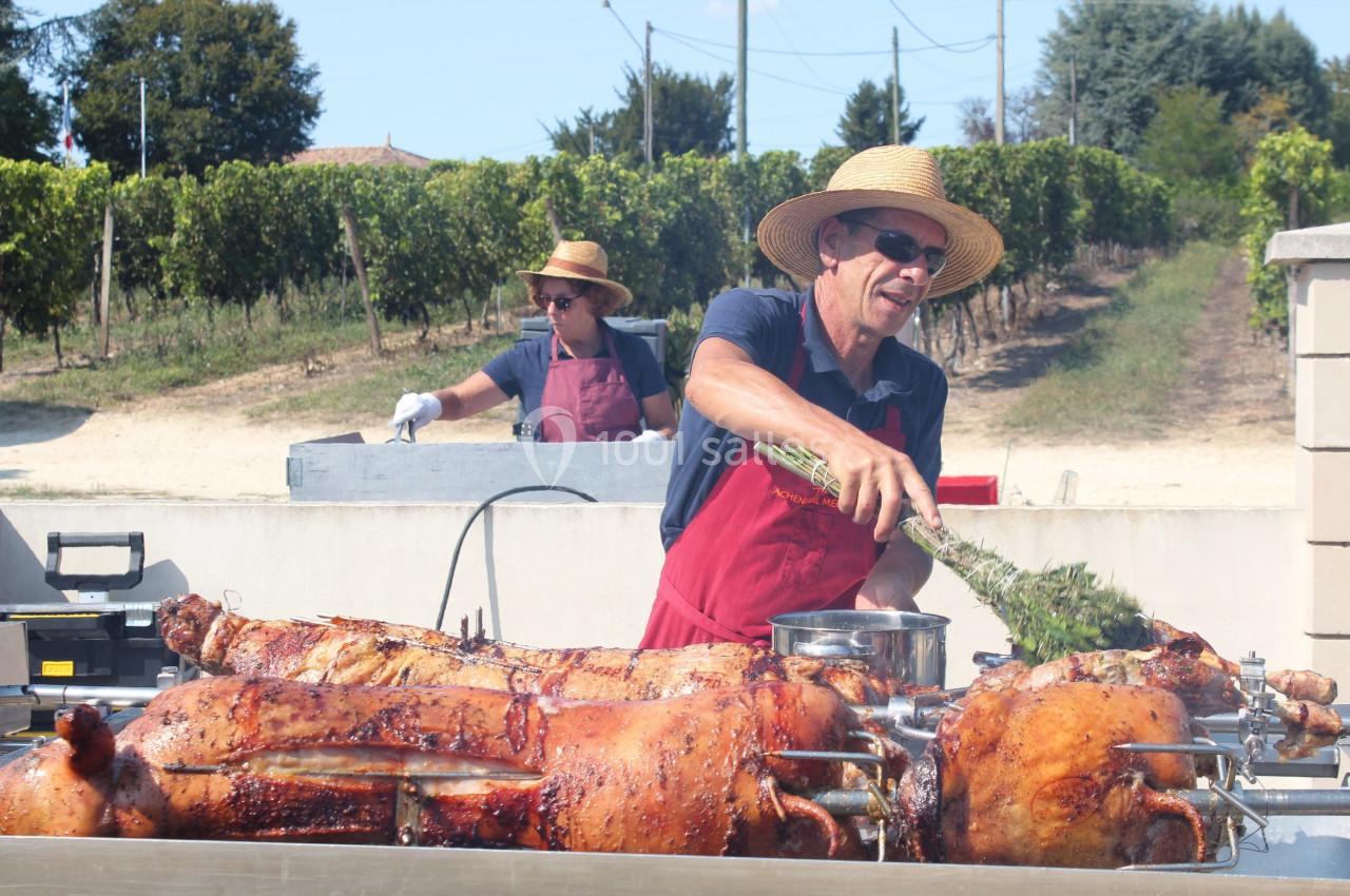 Deux cuisiniers en tablier et chapeau préparent des porcs rôtis sur des broches en extérieur, près de vignes.