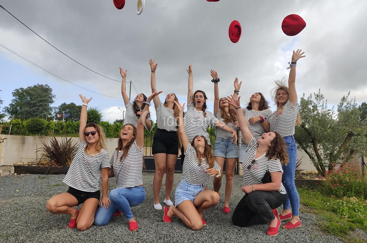 Un groupe de femmes en marinières lance des chapeaux rouges et blancs en l'air dans un espace extérieur gravillonné.