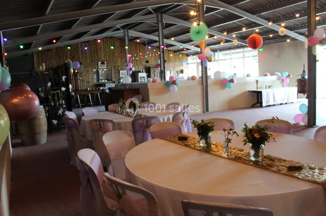 Salle décorée pour un événement avec tables rondes, nappes blanches, guirlandes lumineuses et ballons colorés suspendus.