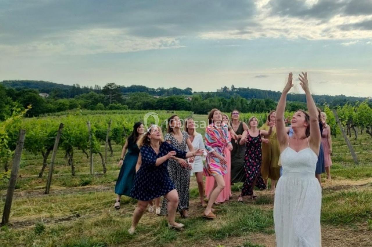 Une mariée lance son bouquet vers un groupe de femmes dans un vignoble sous un ciel partiellement nuageux.