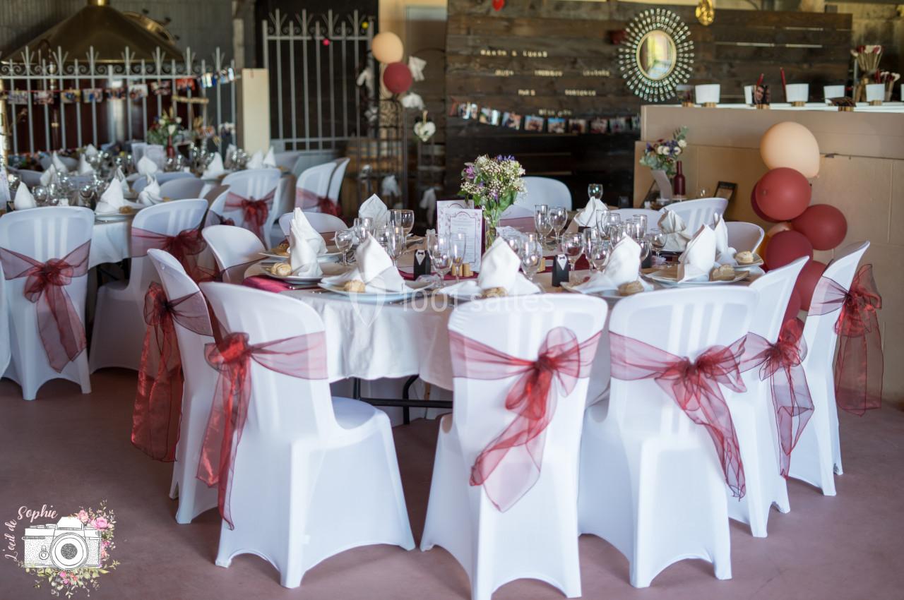 Salle décorée pour un mariage avec des tables rondes, nappes blanches, chaises ornées de rubans rouges et ballons.