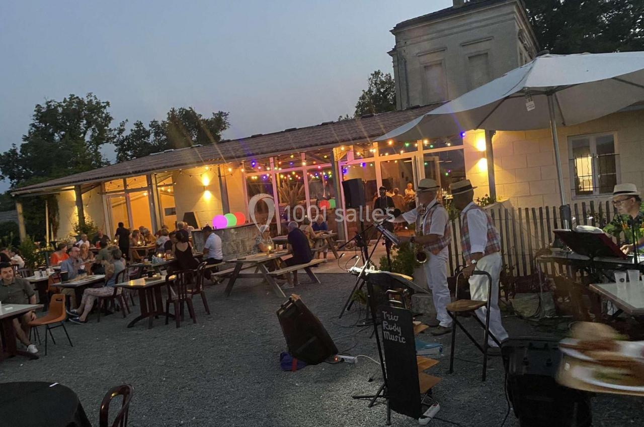 Terrasse animée en soirée avec des convives attablés, un groupe de musiciens jouant et des guirlandes lumineuses.