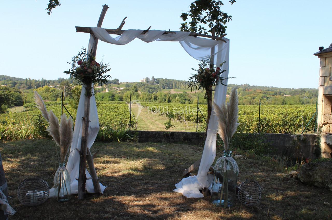 Arche de mariage en bois décorée de voilages blancs et de fleurs, avec vue sur un vignoble sous un ciel dégagé.