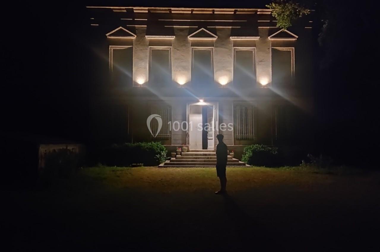 Silhouette d'une personne debout devant une maison éclairée la nuit, entourée de végétation.