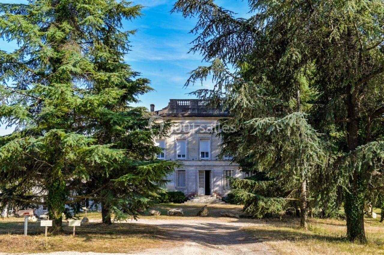 Façade d'une maison en pierre entourée de grands arbres, avec un chemin de terre au premier plan.