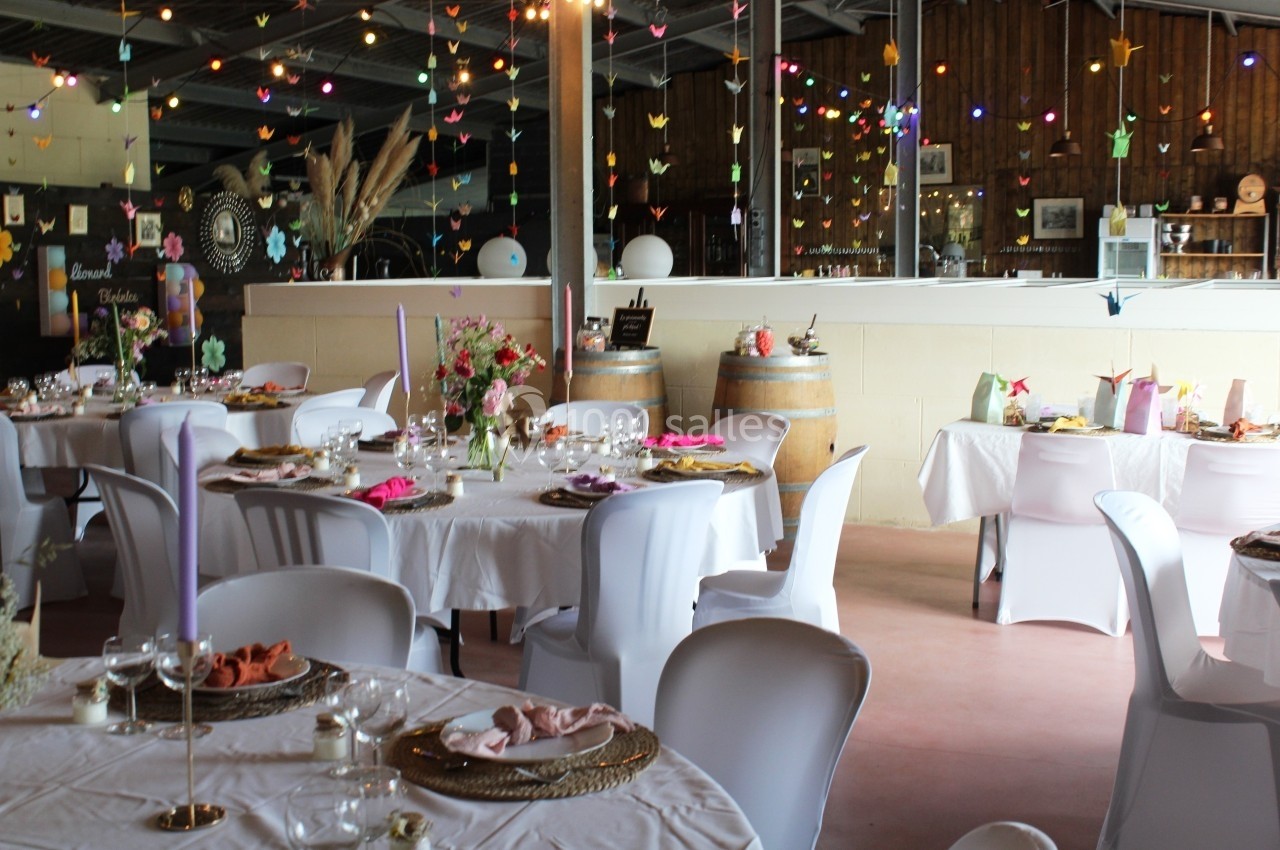 Salle décorée pour un événement avec tables dressées, guirlandes lumineuses et origamis suspendus.
