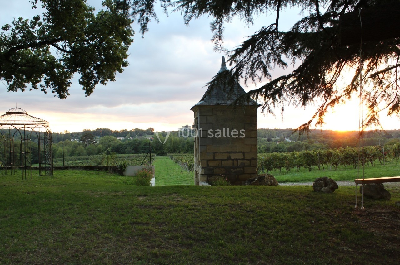Paysage de vignoble au coucher du soleil avec une petite tour en pierre et une pergola métallique sur la gauche.