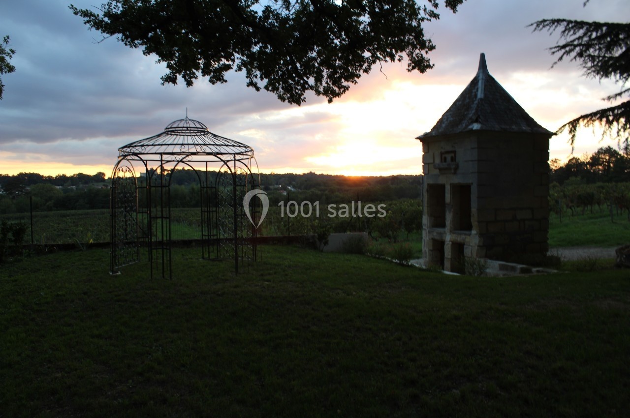 Structure métallique et petite tour en pierre dans un jardin au coucher du soleil, avec des vignes en arrière-plan.