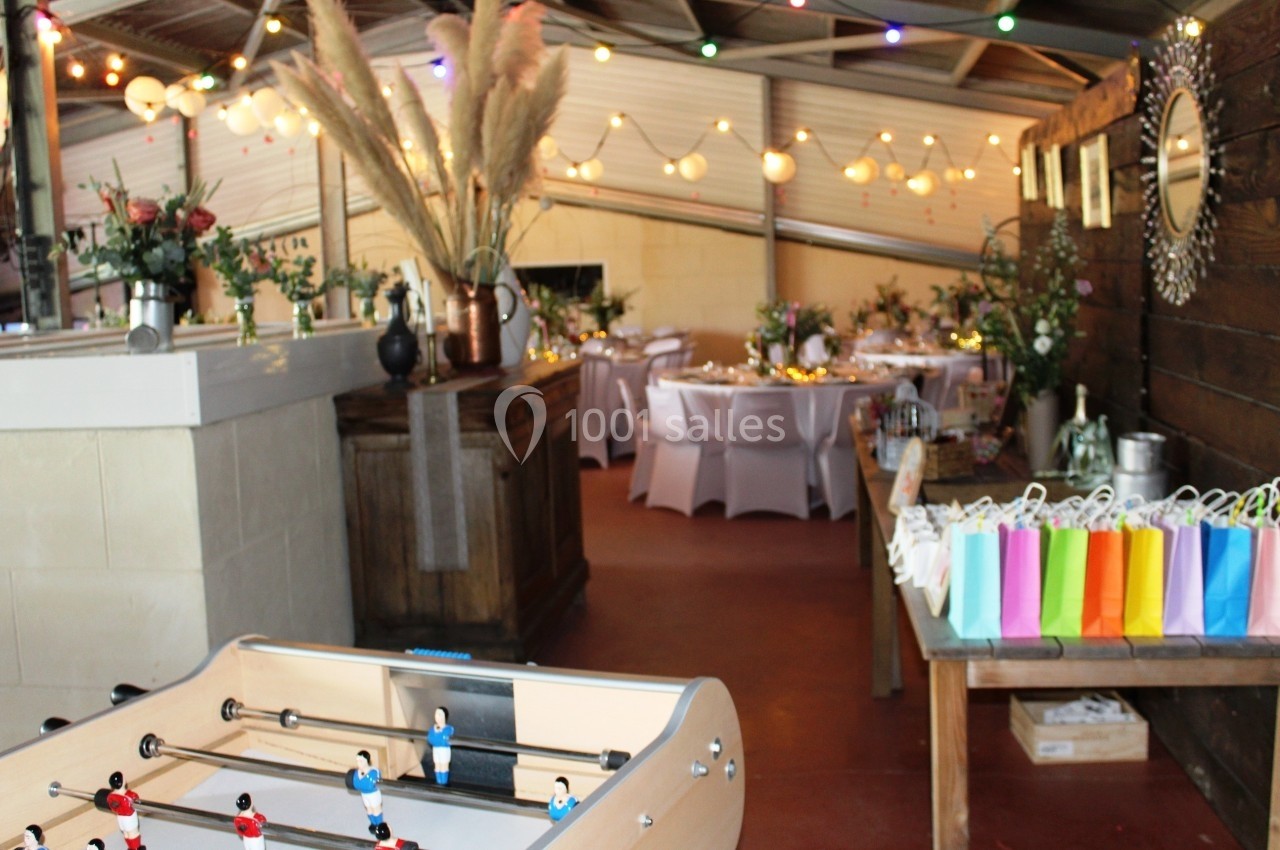 Salle décorée pour un événement avec des tables dressées, des lumières suspendues et un baby-foot au premier plan.