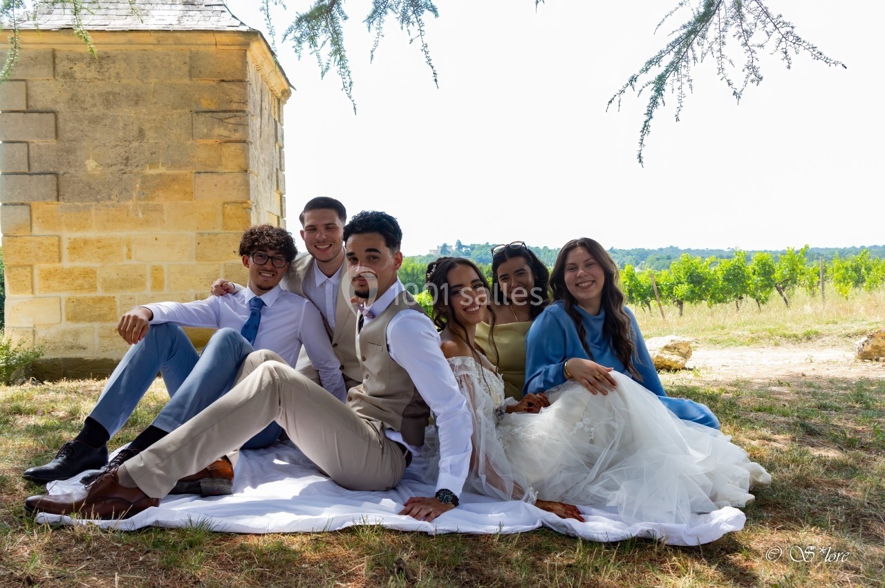 Un groupe de jeunes adultes assis sur une couverture à l'extérieur, près d'un bâtiment en pierre et de vignes en arrière…