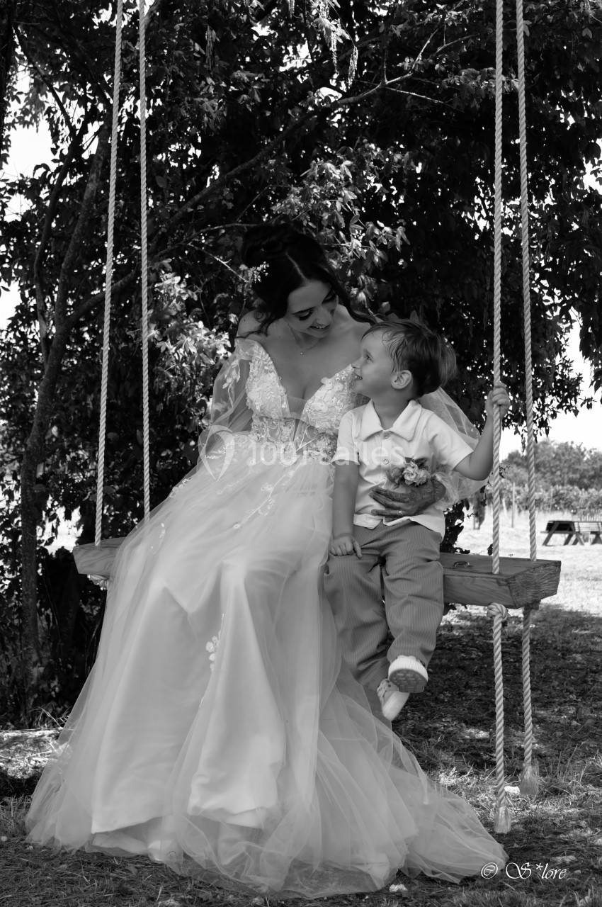 Une femme en robe de mariée sourit à un enfant assis avec elle sur une balançoire, sous un arbre.