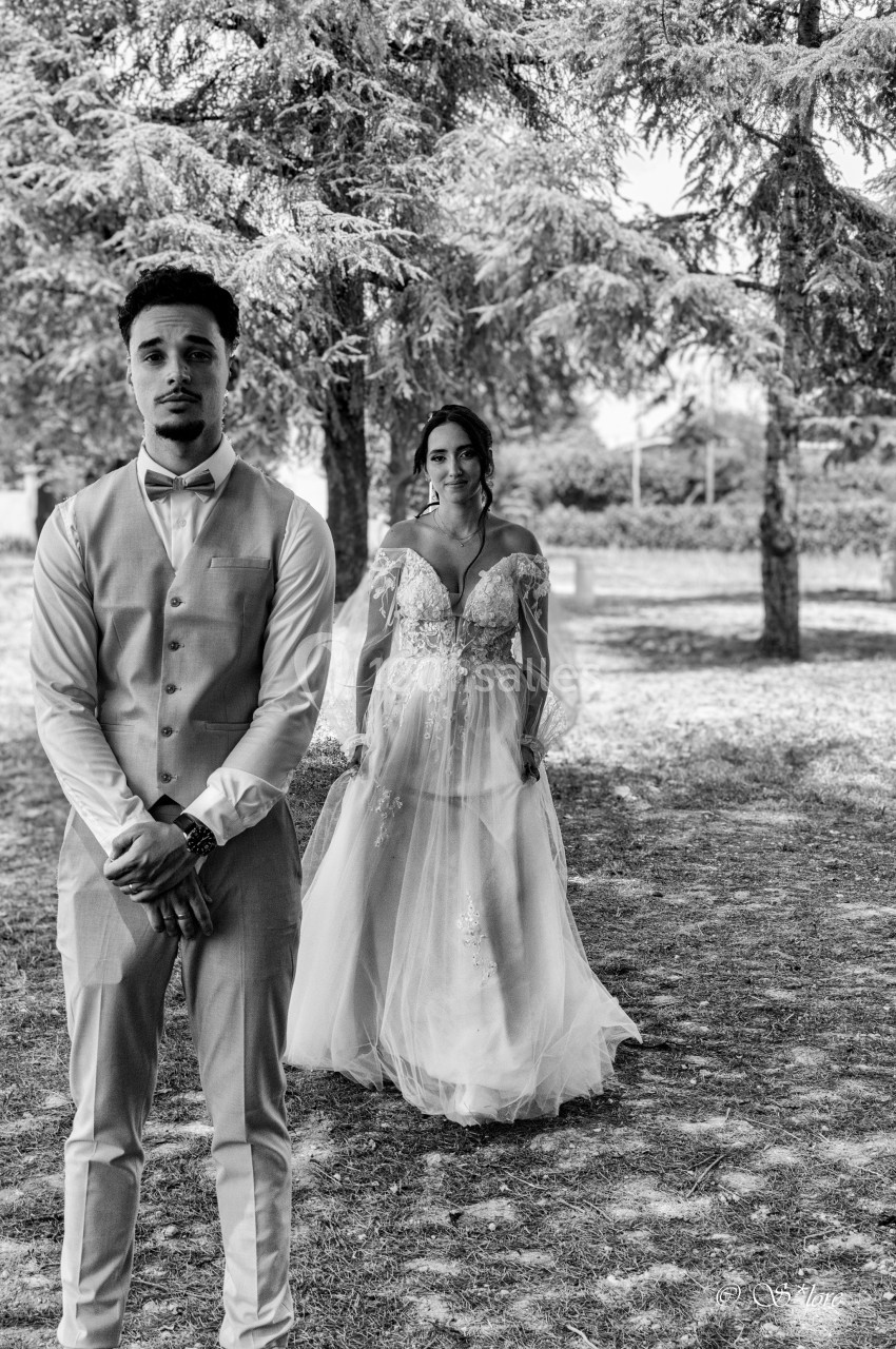 Un homme en gilet et nœud papillon se tient devant une femme en robe de mariée dans un parc arboré.