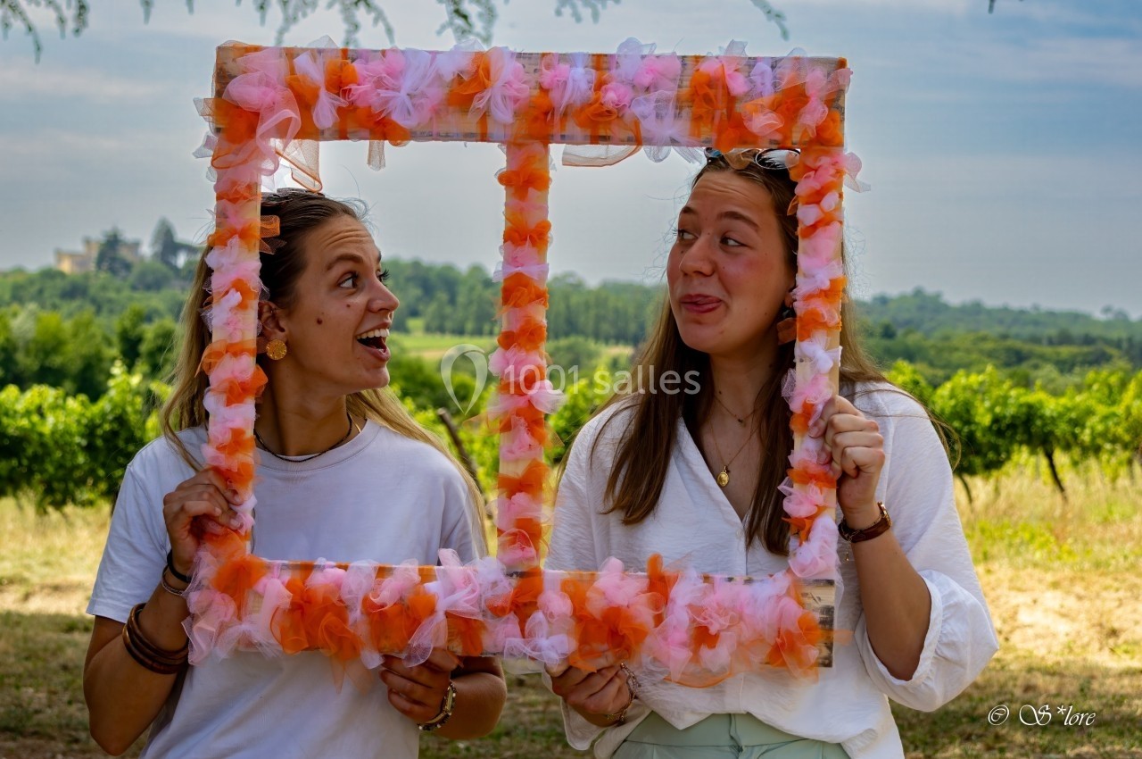 Deux femmes souriantes tiennent un cadre décoré de fleurs orange et blanches dans un paysage champêtre.