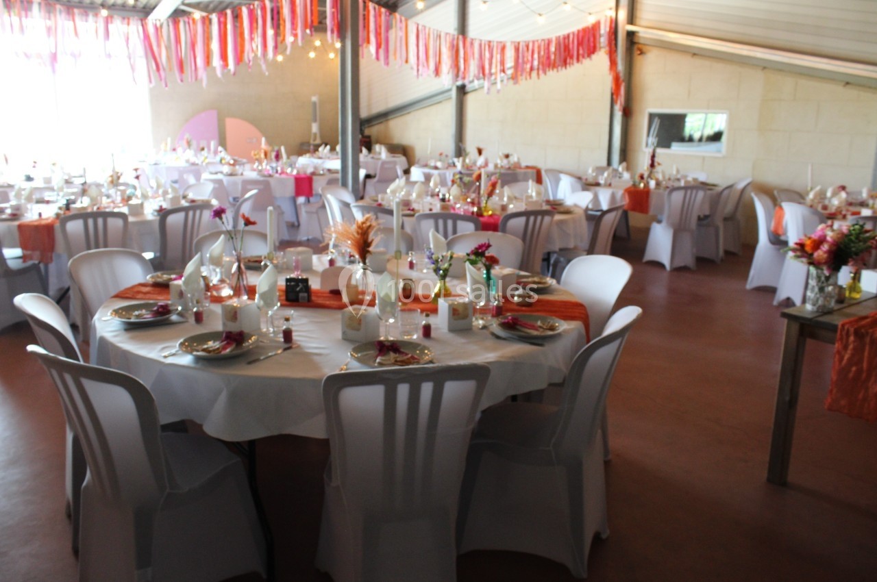Salle décorée pour un événement avec des tables rondes dressées, nappes blanches et décorations colorées suspendues.