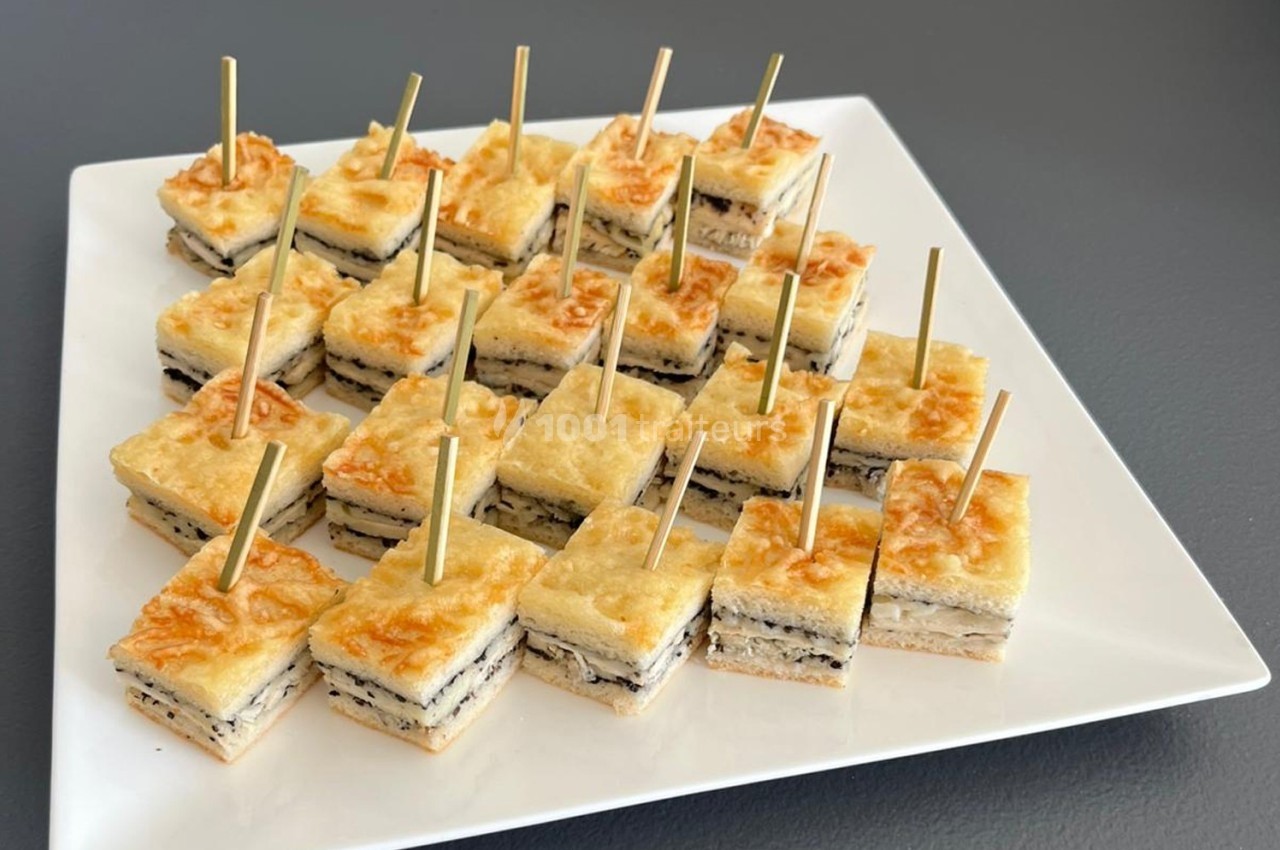 Carrés de croque-monsieur garnis de fromage et truffe, présentés sur un plateau blanc avec des piques en bois.