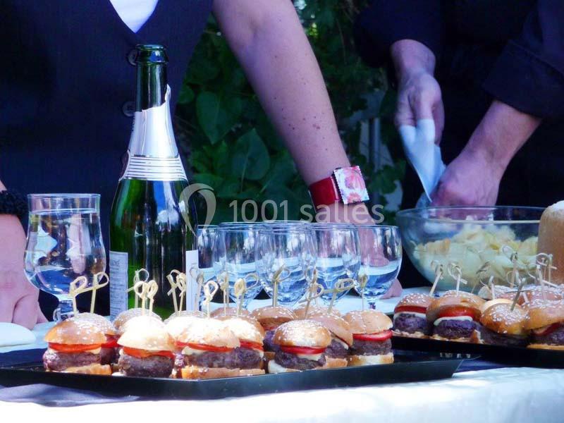Plateau de mini-burgers sur une table avec des verres, une bouteille de champagne et des personnes en arrière-plan.