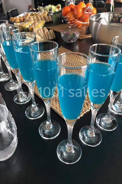 Verres de cocktail bleu alignés sur une table noire, entourés de fruits et de plats en arrière-plan.