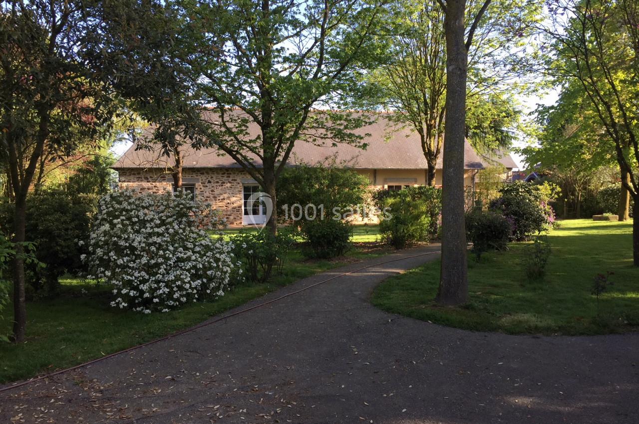 Allée pavée bordée d'arbres et d'arbustes fleuris menant à une maison en pierre sous un toit en ardoise.