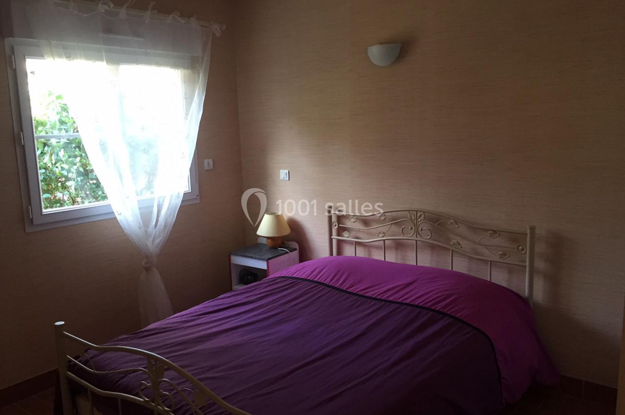 Chambre avec lit double à cadre métallique, draps violets, lampe de chevet et fenêtre avec rideaux blancs.