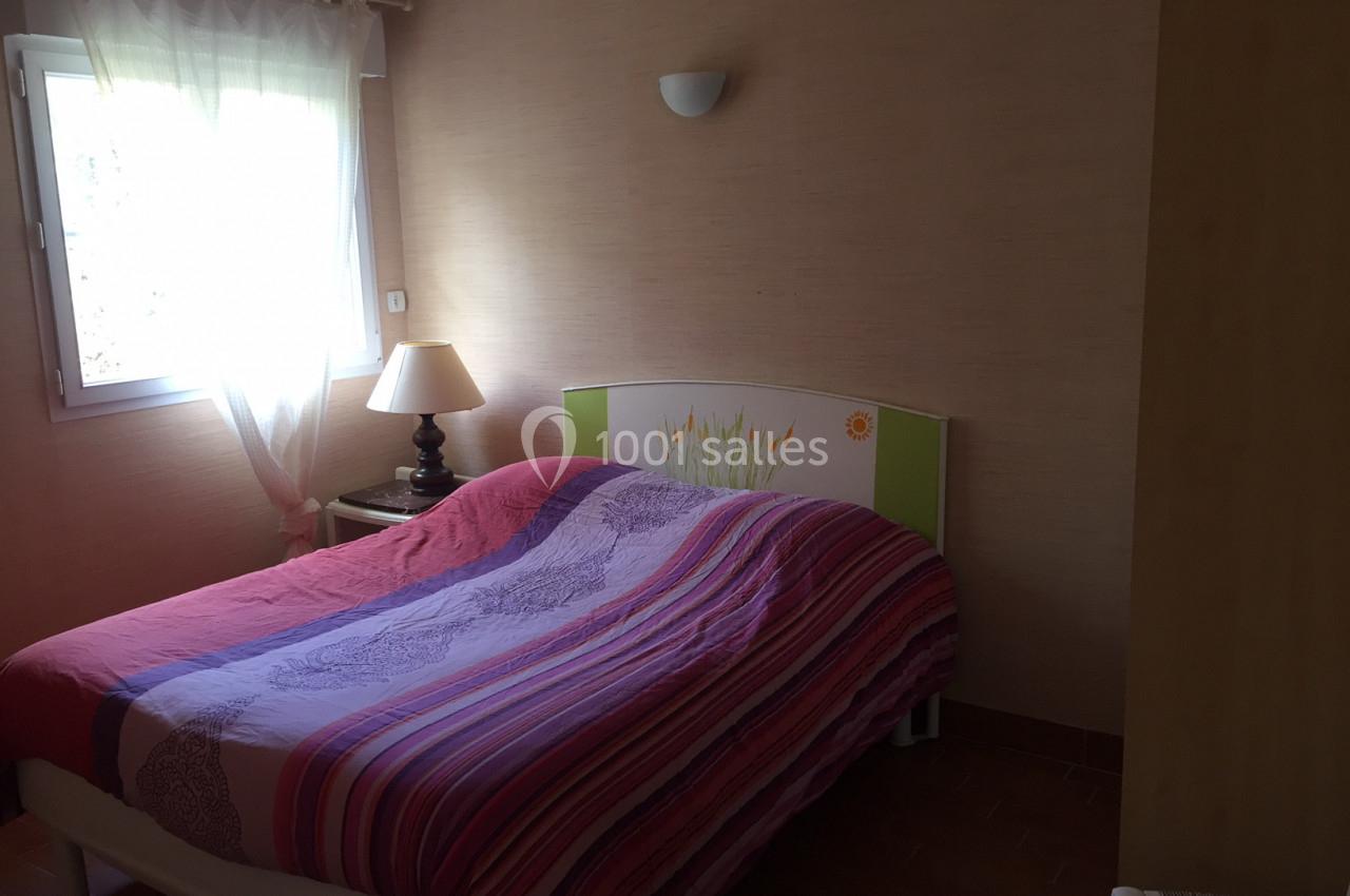 Chambre avec un lit double couvert d'une couette colorée, une lampe de chevet et une fenêtre avec rideaux blancs.