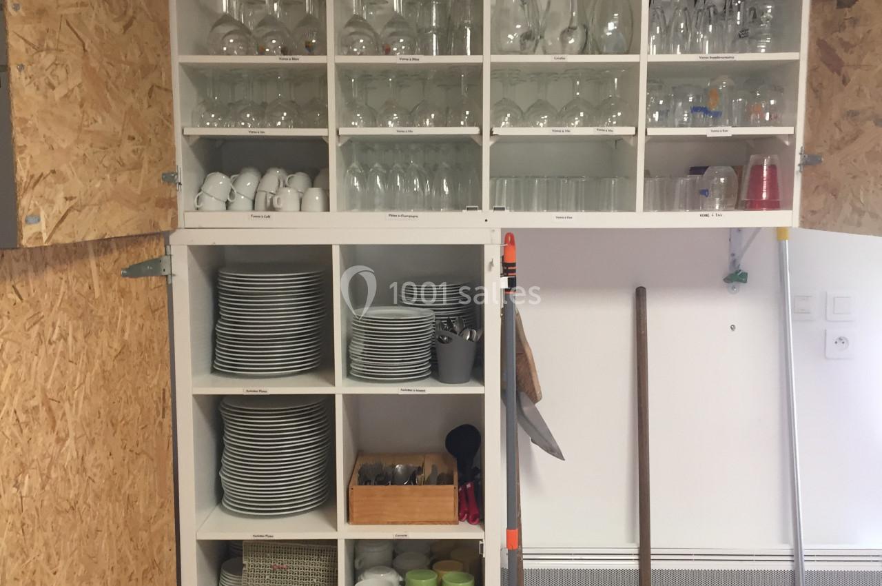 Étagère blanche ouverte contenant des verres, assiettes, tasses et ustensiles, avec balais rangés sur le côté.