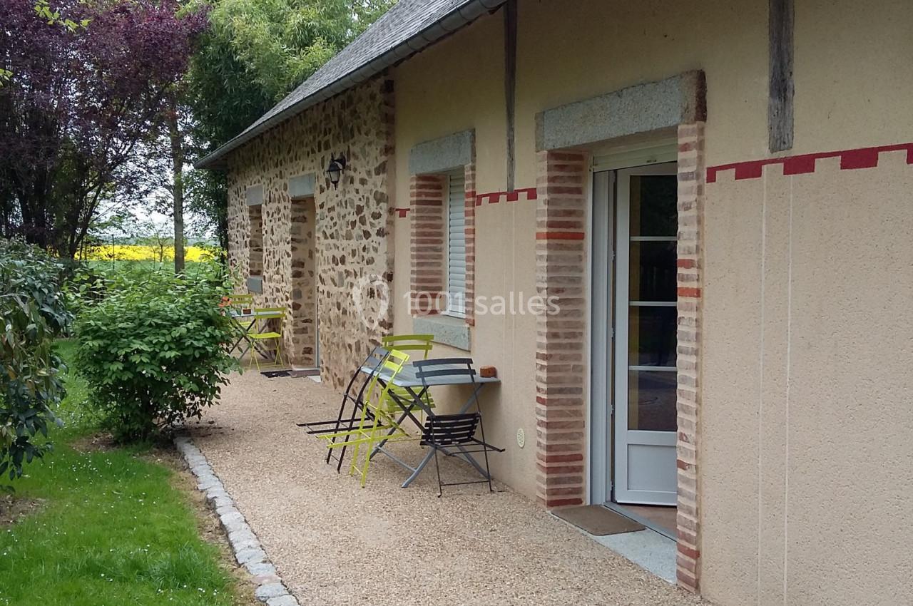 Façade d'une maison en pierre et crépi avec terrasse, mobilier de jardin et allée gravillonnée bordée de verdure.