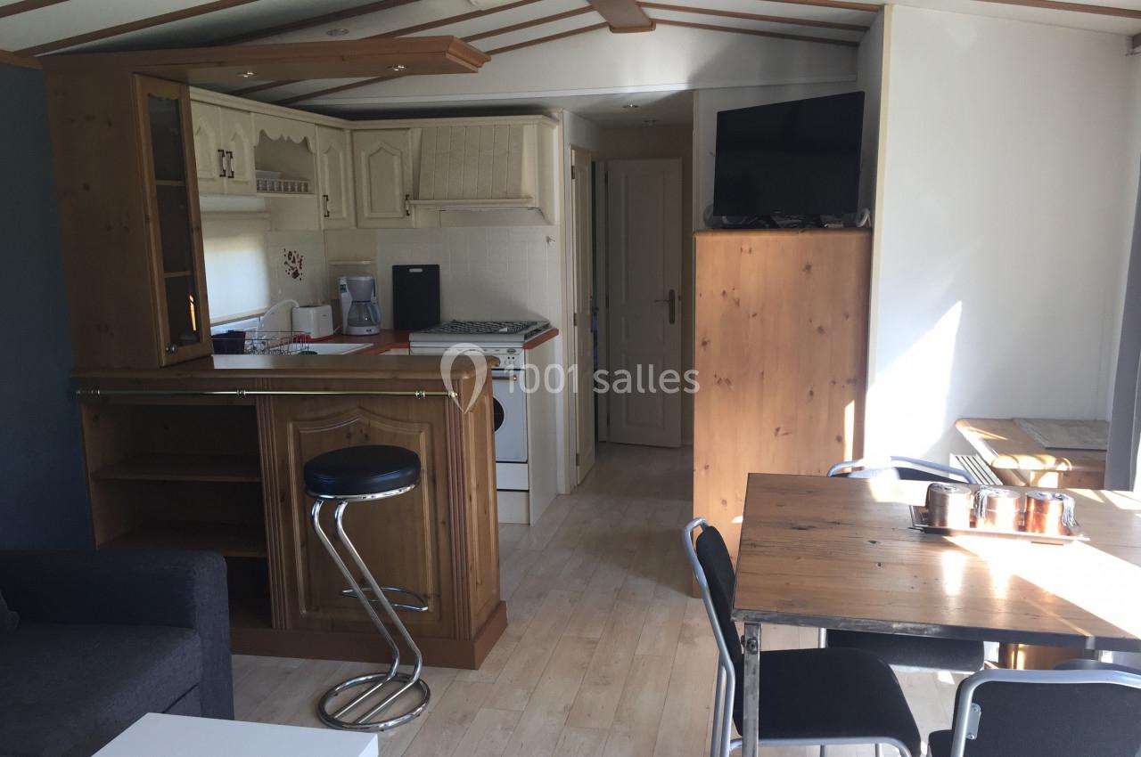 Intérieur d'un mobil-home avec cuisine équipée, table à manger, tabouret de bar et télévision sur un meuble en bois.