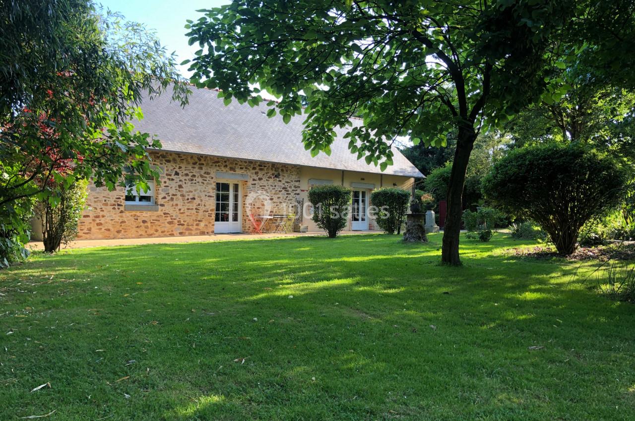 Maison en pierre avec toit en ardoise entourée d'un jardin verdoyant et d'arbres sous une lumière ensoleillée.